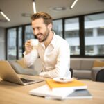 Business man smiling as he holds a mug and uses his laptop at his desk.