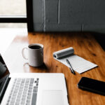 Wood desk with a laptop, cell phone, notepad, pen and a cup of coffee.