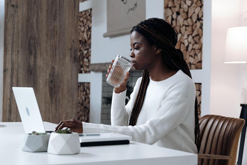 Young woman sitting at her desk and drinking a glass of water as she works on her laptop.