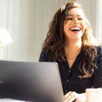 Woman sitting at desk and laughing as she works on her laptop.