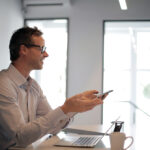 Businessman sitting at desk and smiling as he uses his cellphone and laptop.