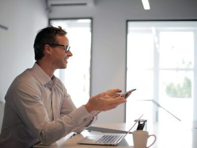 Businessman sitting at desk and smiling as he uses his cellphone and laptop.