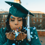 Woman wearing green graduation cap and gown as she blows confetti into the air.