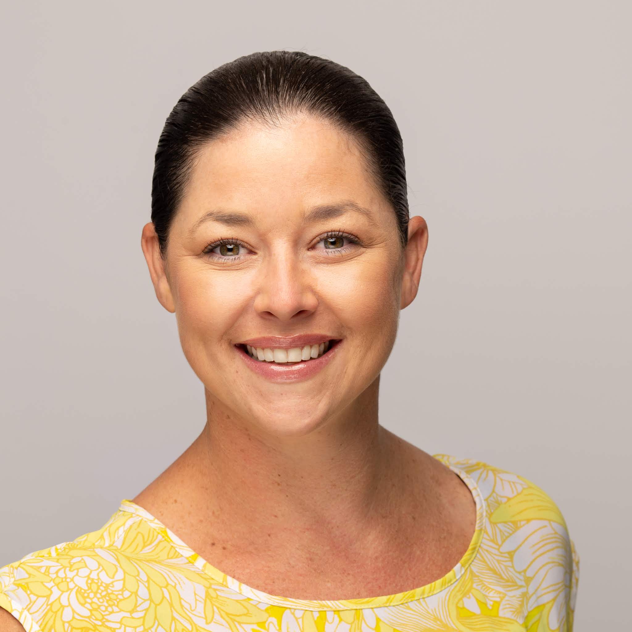 A smiling woman with dark hair pulled back, wearing a yellow and white floral top, poses in front of a plain light background, representing the PC Bennett Solutions team.