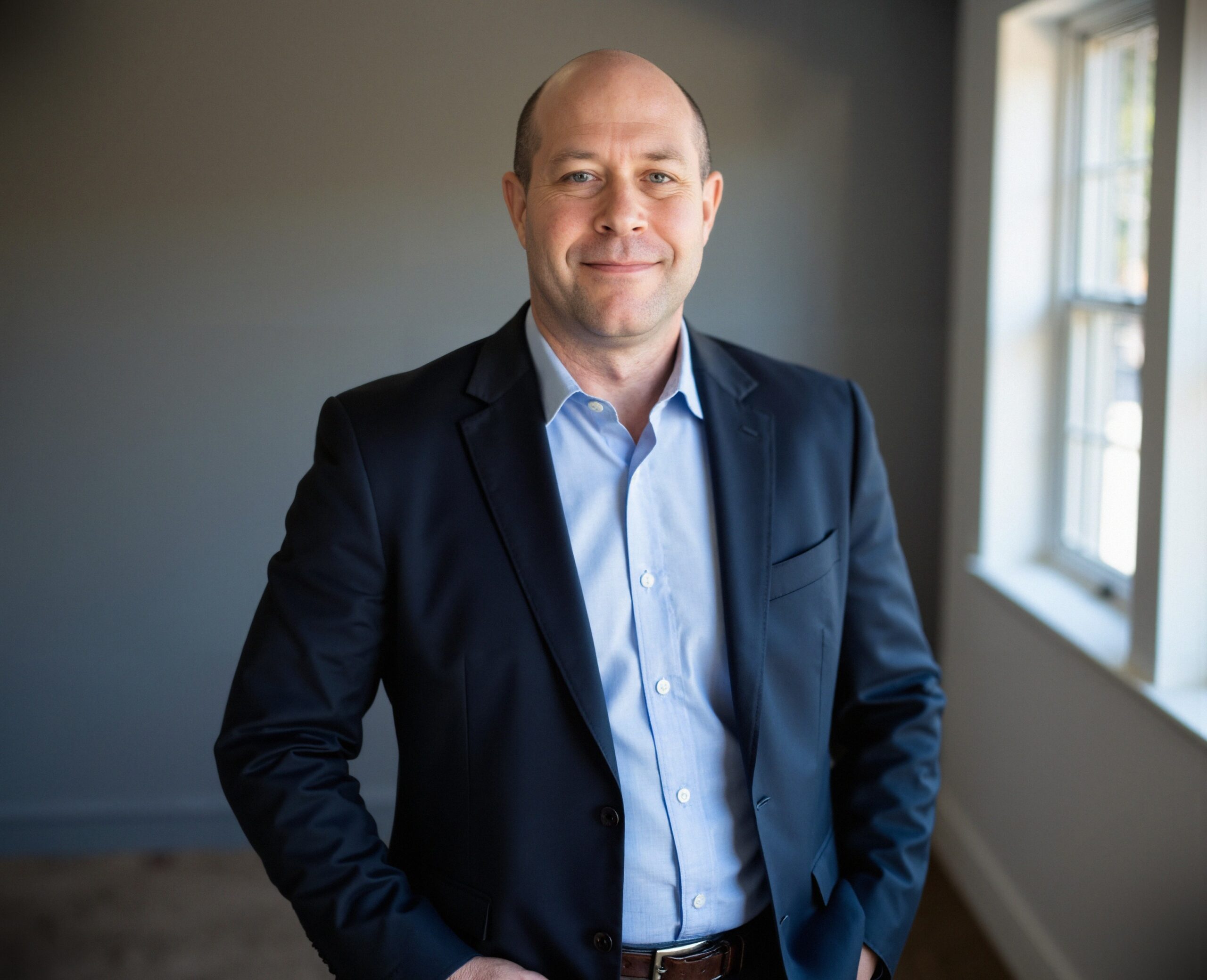 A man in a dark suit and light blue shirt stands indoors, smiling with one hand in his pocket, next to a window letting in natural light, representing the professionalism of the PC Bennett Solutions team.