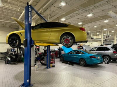 A yellow car is lifted on a hydraulic car lift in an auto repair shop, with its front wheel removed. Several cars and mechanics working are visible in the well-lit, spacious garage.