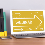A laptop on a wooden desk displays the word "WEBINAR" on its screen with white arrows, hinting at a high performance expo. Next to it are four books held by a black bookend shaped like a person, and a smartphone lies on the desk.