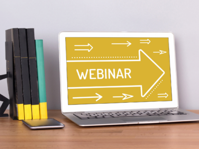 A laptop on a wooden desk displays the word "WEBINAR" on its screen with white arrows, hinting at a high performance expo. Next to it are four books held by a black bookend shaped like a person, and a smartphone lies on the desk.
