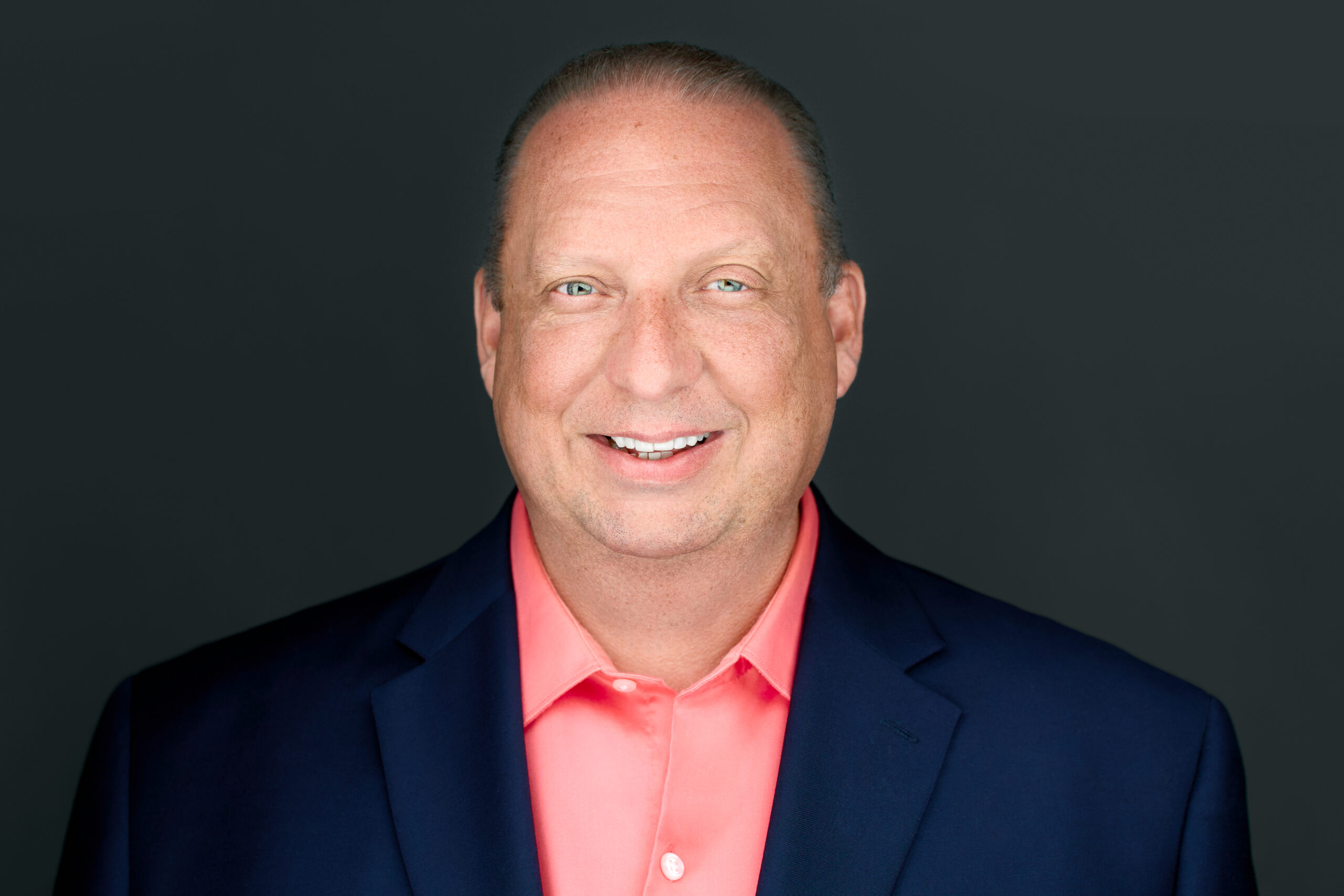 A middle-aged man with short light hair smiles at the camera. He is wearing a dark blue blazer over a bright coral button-up shirt, representing the PC Bennett Solutions team against a plain, dark background.