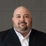 A bald man with a beard, representing the PC Bennett Solutions team, wears a dark suit jacket and white dress shirt, posing in front of a neutral, blurred background with a calm, neutral expression.