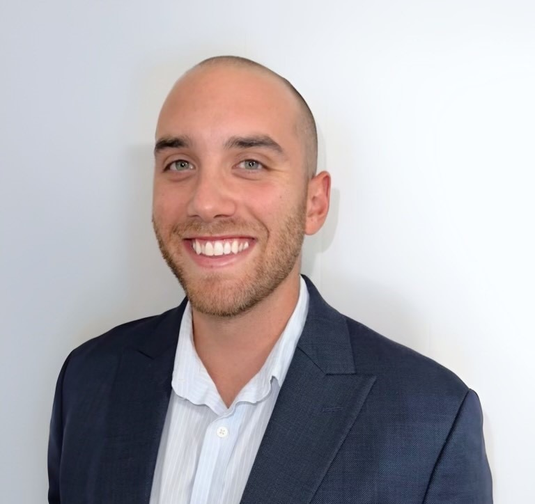 A smiling man with a shaved head and short beard, wearing a dark blazer and light-colored collared shirt, stands against a plain light background—representing the professionalism of the PC Bennett Solutions team.