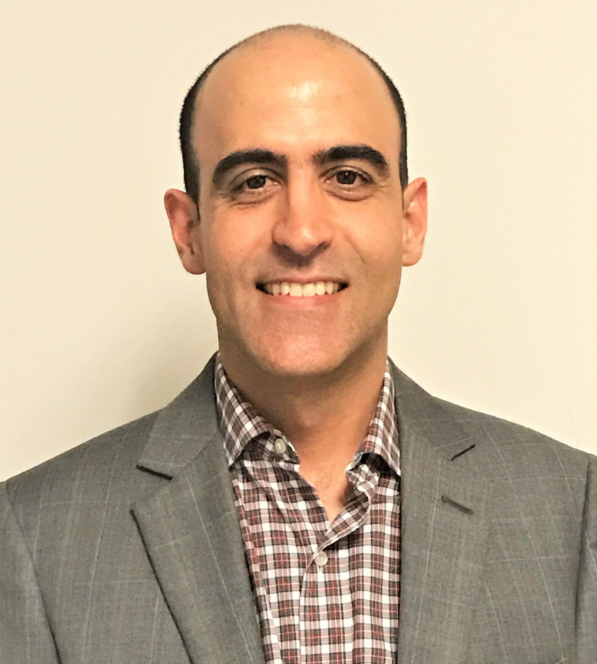 A man with short dark hair and a receding hairline smiles at the camera. He is wearing a gray suit jacket over a red and white checkered shirt, representing the PC Bennett Solutions team against a plain light-colored background.