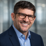 A man with short brown hair, wearing glasses, a blue dress shirt, and a dark blazer, smiles at the camera. The blurred modern office background suggests he is part of the PC Bennett Solutions team.
