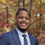 A man in a blue checked suit, white shirt, and navy tie smiles outdoors with the vibrant autumn-colored trees in the background, representing the professional spirit of the PC Bennett Solutions team.