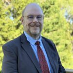 A middle-aged man with glasses, a beard, and a receding hairline wears a dark suit, light blue shirt, and red patterned tie, smiling outdoors with green trees behind him—a proud member of the PC Bennett Solutions team.