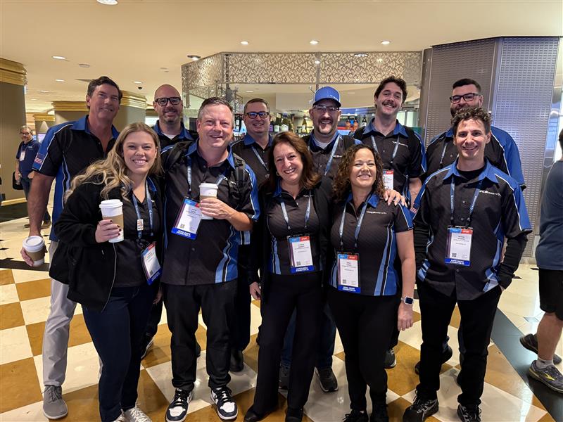 A group of people wearing matching black and blue shirts pose and smile together indoors at an event, with some holding coffee cups and wearing name badges.