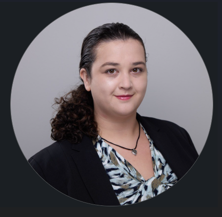 A member of the PC Bennett Solutions team with long curly dark hair pulled back, wearing a black blazer over a patterned blouse and a necklace, poses for a professional headshot against a plain light gray background.