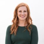 A woman with long, wavy red hair is smiling at the camera. She is wearing a dark green long-sleeve top and stands against a plain white background, representing the friendly professionalism of the PC Bennett Solutions team.