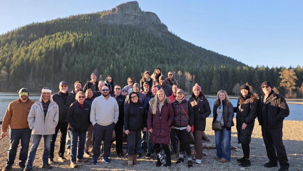 A group of about 30 people pose and smile for a photo outdoors on a sunny day, with a forest and large rocky hill in the background. Most are wearing jackets and sunglasses, suggesting cool weather.