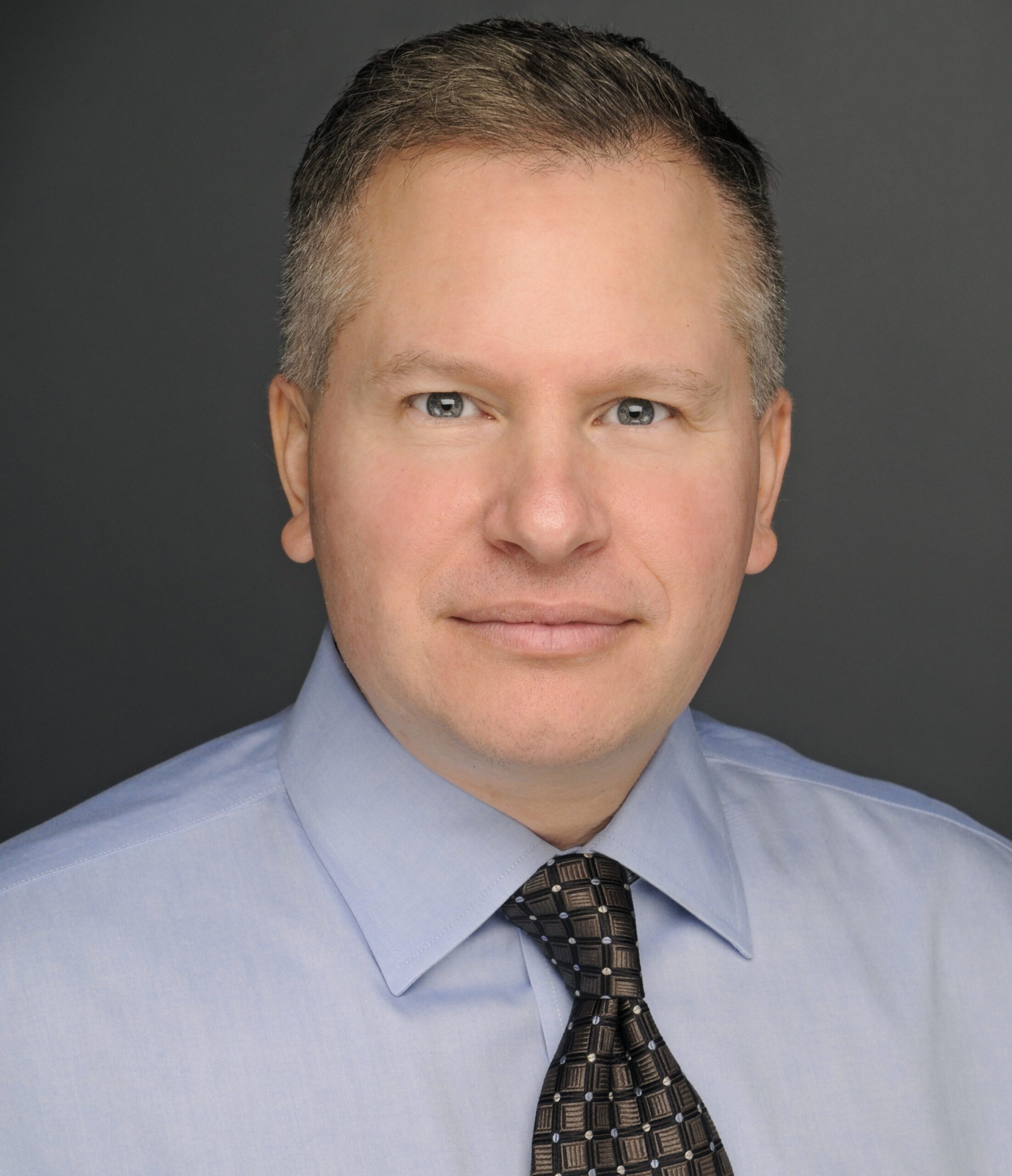 A man with short, light brown hair wearing a light blue dress shirt and a patterned dark tie, representing the PC Bennett Solutions team, poses against a plain dark background.