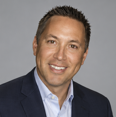 Smiling man with short dark hair, wearing a navy blue suit jacket and light blue shirt, posing in front of a plain gray background—representing the professionalism of the PC Bennett Solutions team.