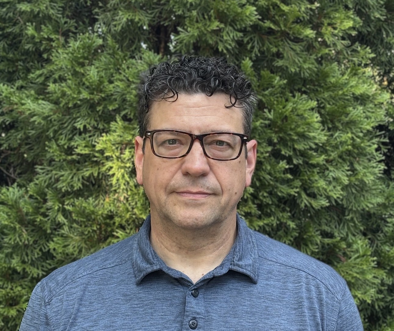 A man with short curly hair and glasses, wearing a blue collared shirt, stands outdoors in front of green leafy bushes, looking directly at the camera with a neutral expression, as if ready to represent the PC Bennett Solutions team.