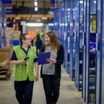 Two people walk through a warehouse aisle, smiling and talking. One wears a green safety vest and holds a device; the other holds a clipboard. Shelves filled with boxes line both sides.