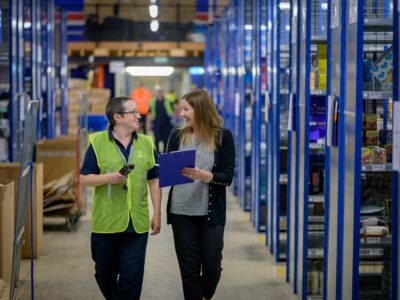 Two people walk through a warehouse aisle, smiling and talking. One wears a green safety vest and holds a device; the other holds a clipboard. Shelves filled with boxes line both sides.